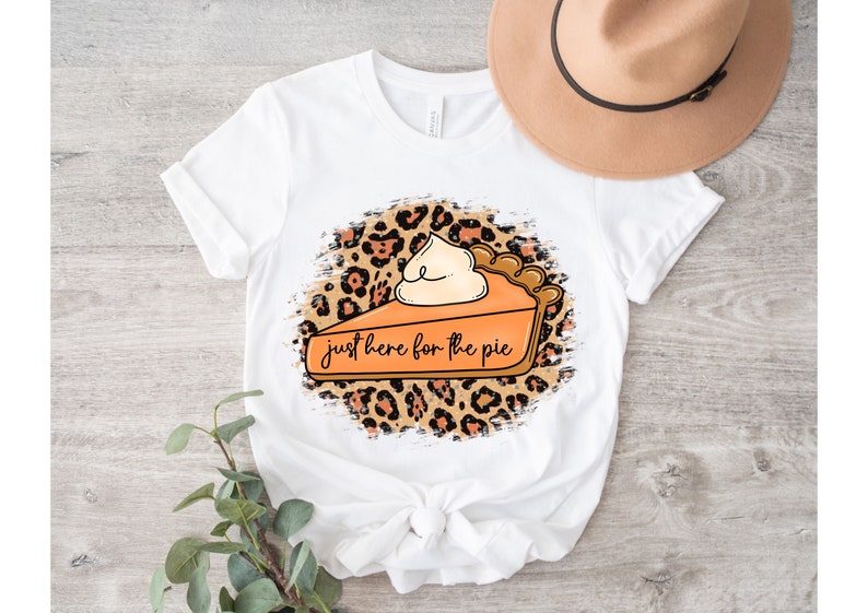 May include: White t-shirt with a graphic of a slice of pumpkin pie with whipped cream on top. The pie is on a leopard print background with the text "just here for the pie".