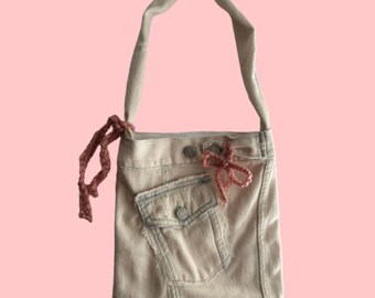 May include: A light beige denim tote bag with a large front pocket and a pink crocheted strap. The bag has a pink crocheted bow on the front.