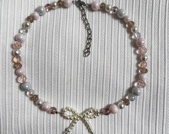 May include: A delicate pearl necklace with a pink and white color scheme. The necklace features a small pearl bow charm.