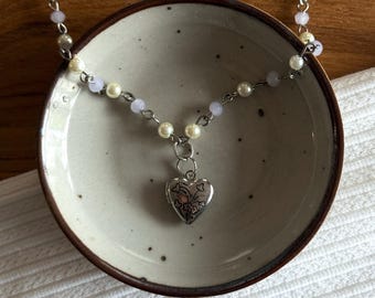 May include: A delicate necklace featuring a silver heart-shaped locket pendant with floral detailing. The necklace is strung with small, creamy pearls and pale lavender beads. The necklace is displayed in a small, speckled ceramic bowl.