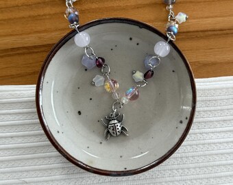 May include: A delicate necklace featuring a silver bug pendant and a chain adorned with various beads in shades of purple, white, and iridescent colors. The necklace is displayed on a small, round, speckled ceramic dish.