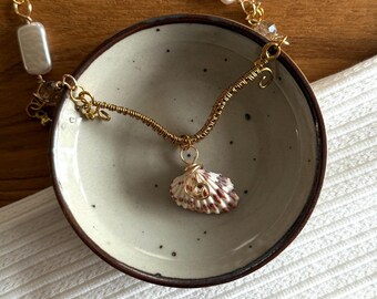 May include: A handcrafted necklace featuring a seashell pendant. The pendant is a small, white and brown seashell, wrapped in gold-colored wire. The necklace chain includes rectangular white beads and gold-colored wire links. The necklace is displayed in a small, round, ceramic bowl.