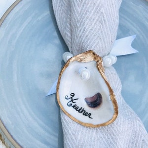 May include: A white napkin with a gold oyster shell place card. The oyster shell has a black and white illustration of an oyster and the name "Heather" written in black cursive.
