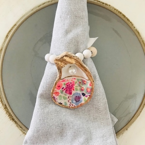 May include: A gray linen napkin with a white and gold oyster shell napkin ring. The oyster shell has a floral pattern in pink, blue, and green colors.