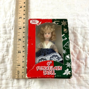 May include: A 4 inch porcelain doll ornament in a red, white, and green box. The doll is wearing a white blouse with a blue skirt and white lace trim. The box has the text "4" inch porcelain doll" on it.