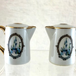 May include: Two white ceramic salt and pepper shakers with gold trim and a Disneyland logo featuring a depiction of Cinderella's Castle.