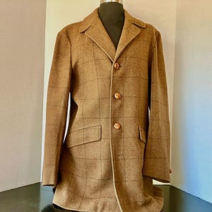 May include: A brown tweed jacket with a plaid pattern. The jacket has a notched collar, three buttons, and two front pockets. The jacket is displayed on a mannequin.