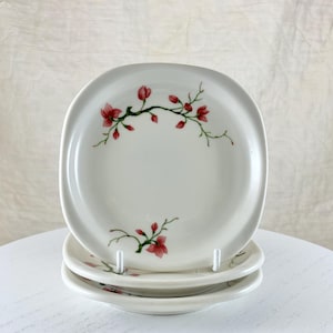May include: A stack of four white, square-shaped plates with rounded corners. Each plate is decorated with a floral design of red flowers and green stems. The plates are stacked on a white surface.