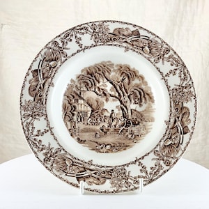 May include: A white ceramic plate with a brown transferware design. The center features a rural scene with a house, trees, and figures. The rim has a floral and scroll border. A decorative piece for serving or display.