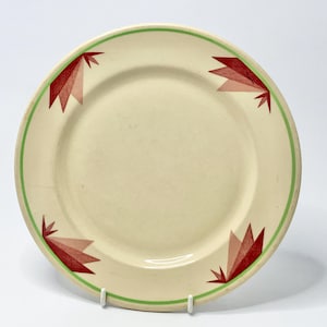 May include: Cream-colored ceramic plate with a green border and red geometric designs. The plate features a series of stylized red and pink shapes around the rim. A vintage-style plate for serving food.