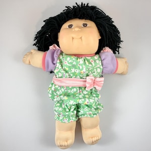 May include: A vintage doll with black yarn hair, brown eyes, and a soft, rounded face. The doll wears a green floral print outfit with pink and purple accents, and a pink bow. The doll's arms and legs are extended, and it is set against a white background.