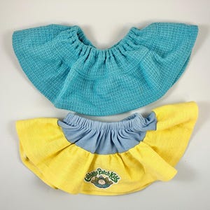 May include: Two doll skirts. One is turquoise blue with a waffle weave texture. The other is yellow with a light blue waistband and a ruffled hem. The Cabbage Patch Kids logo is on the yellow skirt.