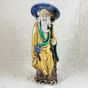May include: A ceramic figurine of a man with a long white beard, wearing a blue hat and a yellow robe with green accents. He is holding a staff in his right hand and a blue and white object in his left hand.