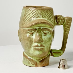 May include: A ceramic mug shaped like a man's head, with a golf club handle. The mug is primarily green and brown, with a textured hat and face. A silver thimble sits next to the mug.