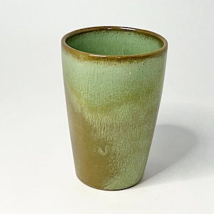 May include: A handmade ceramic tumbler with a green and brown glaze. The cylindrical cup has a slightly flared rim and a smooth, glossy finish. The color transitions from a light green at the top to a darker brown at the base.