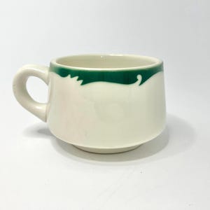 May include: A cream-colored ceramic mug featuring a wide, dark green band around the upper rim. The mug has a rounded body, a short base, and a curved handle on the left side. The surface is smooth and glossy, with a simple, clean design.
