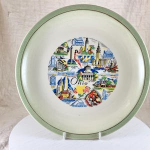 May include: A round, off-white plate with a light green rim. The center features a colorful illustration of Ohio landmarks and the word "Ohio". The plate is displayed on a white stand.