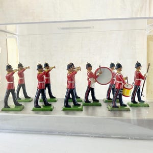 May include: A collection of miniature toy soldiers in a marching band formation, displayed in a clear acrylic case. The soldiers wear red jackets, black pants, and black helmets. Some play trumpets, drums, and carry batons.