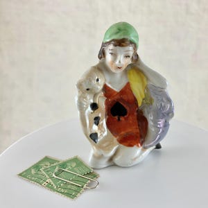 May include: A porcelain figurine of a woman wearing a green hat and a red dress with a black spade on the front. She is holding a small porcelain dog in her arms.