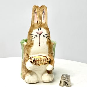 May include: A ceramic bunny figurine with a basket, painted in brown, white, and green. The bunny is holding a basket and has a smiling face. A silver thimble sits next to the figurine.