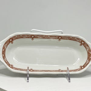 May include: A white, oval-shaped serving dish with a decorative brown border. The dish has a scalloped edge and is displayed on a white surface. The brown border features a repeating floral design.