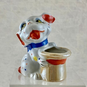 May include: A white ceramic dog figurine with red, blue, and yellow painted details. The dog is wearing a blue bow tie and holding a brown and red painted hat. The hat has a white interior and is designed to be a planter.