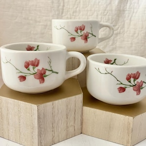 May include: Three white ceramic mugs with handles, each adorned with a delicate floral design featuring pink blossoms and green stems. The mugs are arranged on a gold and wood display, showcasing a charming, handcrafted aesthetic.