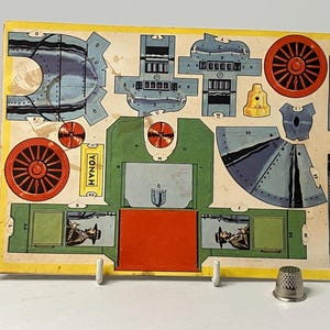 May include: Vintage paper train model kit with cut-out pieces in various shapes and colors, including red wheels, blue panels, and green sections. The word "YONAH" is printed on the side. A silver thimble rests on the surface.