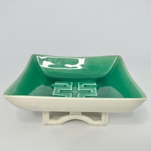 May include: A square ceramic dish with a glossy emerald green interior and a matte white exterior. The center of the dish features a raised geometric Greek key pattern. The dish is supported by a white, open-frame pedestal base with curved feet.