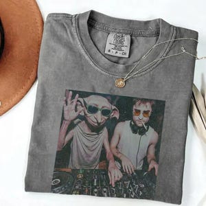 May include: Grey Comfort Colors t-shirt with a graphic of two figures, one like Dobby from Harry Potter and the other like Harry Potter, DJing. The shirt has a crew neck and a small tag. A silver necklace with a pendant is draped on the shirt.