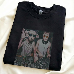 May include: Black crewneck sweatshirt featuring a graphic of two figures with sunglasses and a DJ mixing board. One figure resembles a house elf, the other a man with headphones and a cigarette. The sweatshirt is made of a soft material.