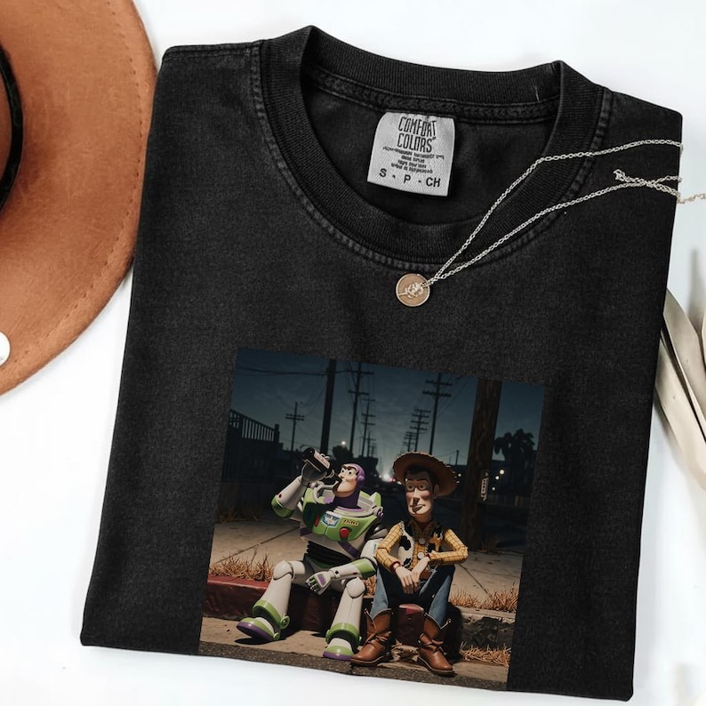 Puede incluir: Una camiseta negra de cuello redondo con un estampado gr&aacute;fico de dos personajes animados sentados en un bordillo por la noche. La camiseta tiene una etiqueta de la marca Comfort Colors en el interior del cuello con el texto Comfort Colors y S P CH.