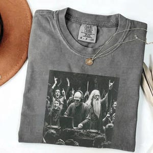 May include: Gray Comfort Colors t-shirt with a black and white graphic of a DJ and a crowd. The shirt has a crew neck and a small tag at the neckline. A silver necklace with a pendant is draped across the shirt.