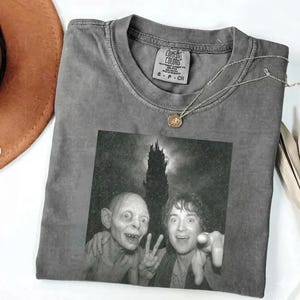 May include: Gray Comfort Colors t-shirt featuring a black and white image of Gollum and Frodo from Lord of the Rings. A silver necklace with a pendant is draped on the shirt. The shirt has a small Comfort Colors tag.