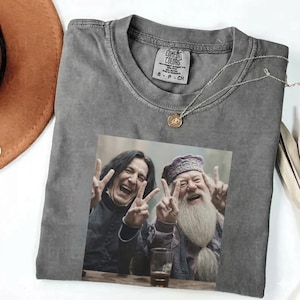 Puede incluir: Camiseta gris Comfort Colors con un estampado de dos personas haciendo el signo de la paz. Un collar plateado con un colgante descansa sobre la camiseta. Un sombrero marrón es visible en la esquina superior izquierda.