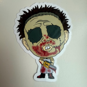 May include: A cartoon sticker of a character with a stitched-up face, dark eyes, and blood splatter. The character is holding a yellow and silver chainsaw and wearing a white apron and black boots. The sticker has a white border.