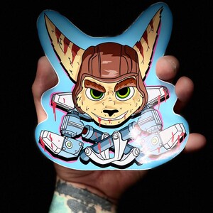 May include: A die-cut sticker featuring a cartoon character with a brown helmet, green eyes, and a mischievous grin. The character is set against a light blue background with stylized mechanical elements and red accents. The sticker is held against a black background.
