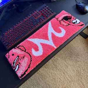 May include: A pink rectangular rug featuring a white "M" design and cartoon character faces at each end. The rug is placed on a black desk next to a red-lit keyboard and a computer monitor.