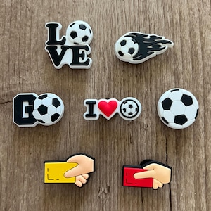 May include: A collection of soccer-themed charms in black, white, and red. Designs include the word "LOVE" with a soccer ball, a soccer ball with flames, the word "GO" with a soccer ball, "I ❤️ soccer", a soccer ball, and hands holding yellow and red cards.