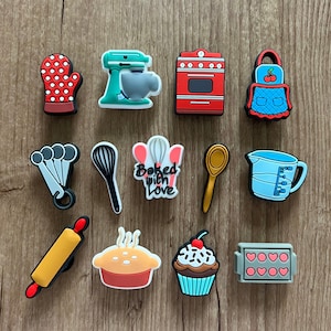 May include: A collection of colorful baking-themed charms, including a red oven, a mixer, a cupcake, and a rolling pin. Other charms include a measuring cup, a whisk, and a mitt. The phrase "Baked with Love" is also featured.