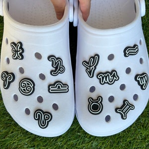 May include: A pair of white clogs adorned with black and white zodiac symbol charms. The charms are placed on the top of the shoes, with various astrological signs visible, including Aries, Taurus, and Gemini.