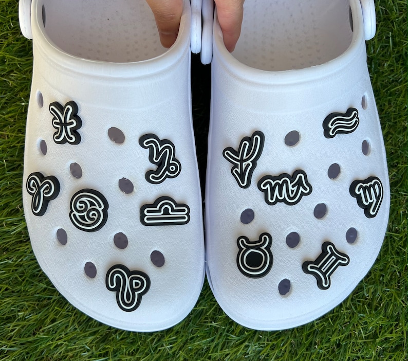 May include: A pair of white clogs adorned with black and white zodiac symbol charms. The charms are placed on the top of the shoes, with various astrological signs visible, including Aries, Taurus, and Gemini.