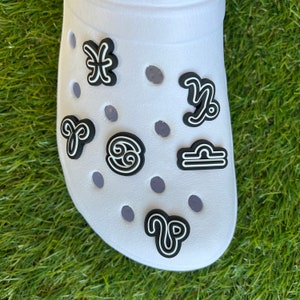 May include: White shoe with several black and white zodiac sign charms. The charms are in the shape of the astrological symbols for Pisces, Aries, Cancer, Capricorn, and Libra. The shoe is on a green grassy surface.