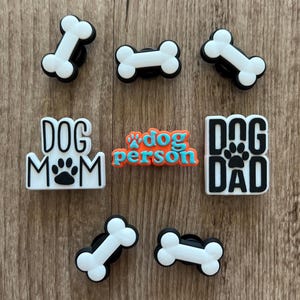May include: A collection of dog-themed charms and accessories. The image features white bone-shaped charms with black borders, along with charms that read "DOG MOM", "dog person", and "DOG DAD". The charms are in various shapes and sizes.