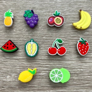 May include: A collection of colorful fruit-shaped charms, including a pineapple, grapes, passion fruit, banana, watermelon, durian, cherries, strawberry, lemon, and lime. Each charm has a white border and is set against a wood-grain background.