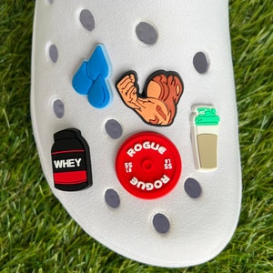 May include: A white shoe adorned with various fitness-themed charms. The charms include a blue water droplet, a flexing arm, a protein powder container with "WHEY" text, a red weight plate with "ROGUE" text, and a shaker cup.