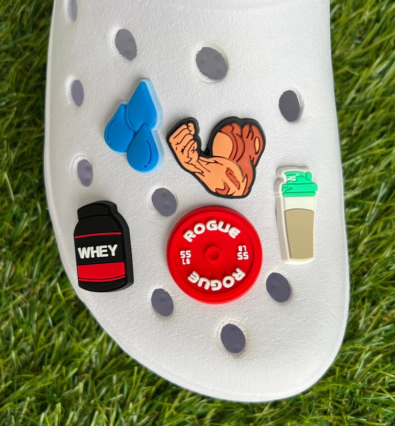 May include: A white shoe adorned with various fitness-themed charms. The charms include a blue water droplet, a flexing arm, a protein powder container with "WHEY" text, a red weight plate with "ROGUE" text, and a shaker cup.