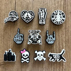 May include: A collection of black and white shoe charms with various designs. The charms include a heart with skeletons, a coffin with skeletons, a witch logo, a hand with the text "KNOW YOUR POWER", a middle finger skeleton hand, and a cat with the text "BLACK MAGIC".