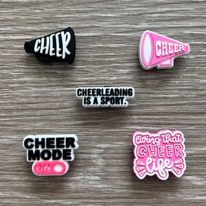 May include: An assortment of cheerleading-themed charms. The charms include a black megaphone with the word "CHEER" in white, a pink megaphone with "CHEER", a charm that says "CHEERLEADING IS A SPORT", a charm that says "CHEER MODE ON", and a charm that says "living that CHEER life".