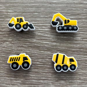 May include: Four colorful shoe charms featuring construction vehicles. The set includes a yellow bulldozer, excavator, dump truck, and cement mixer. Each charm has a black and white outline, and is designed to be attached to shoes.